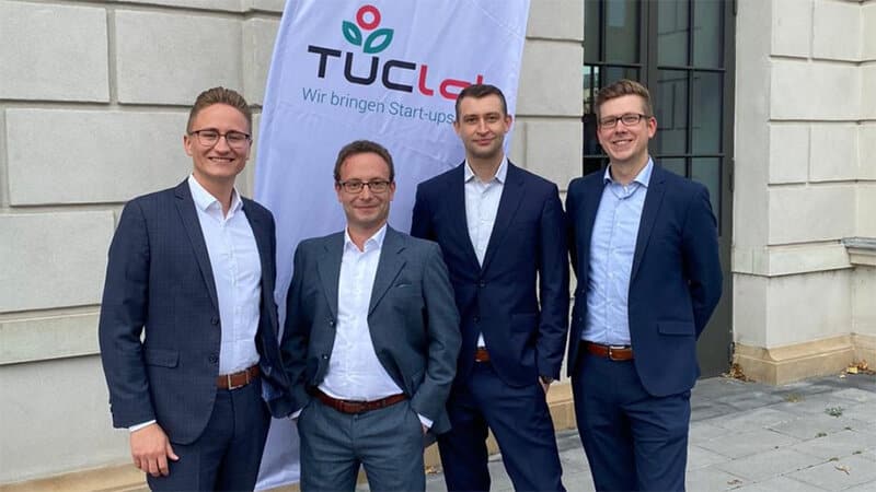 Das vierköpfige Team der plasmotion GmbH steht in Siegerpose vor dem Banner des TUClab Gründer-wettbewerbs